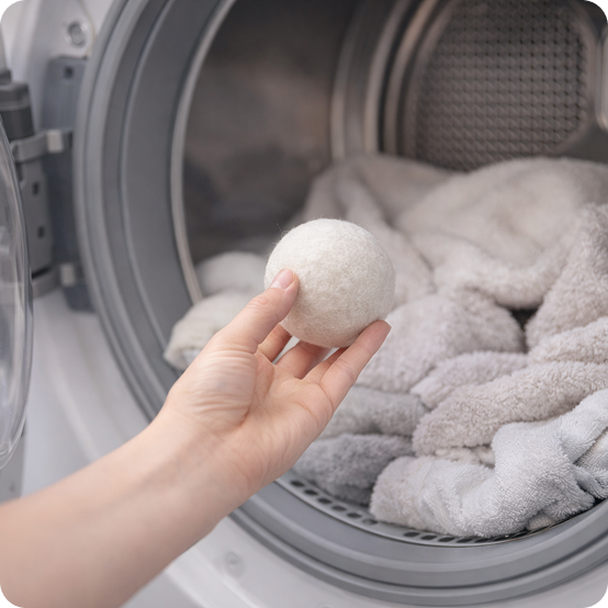 Hand holding a white laundry detergent ball in front of an open dryer with clothes inside.