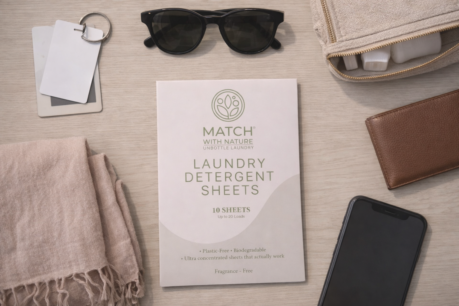 Laundry detergent sheets packaging with a phone, wallet, sunglasses, and towel on a wooden surface.